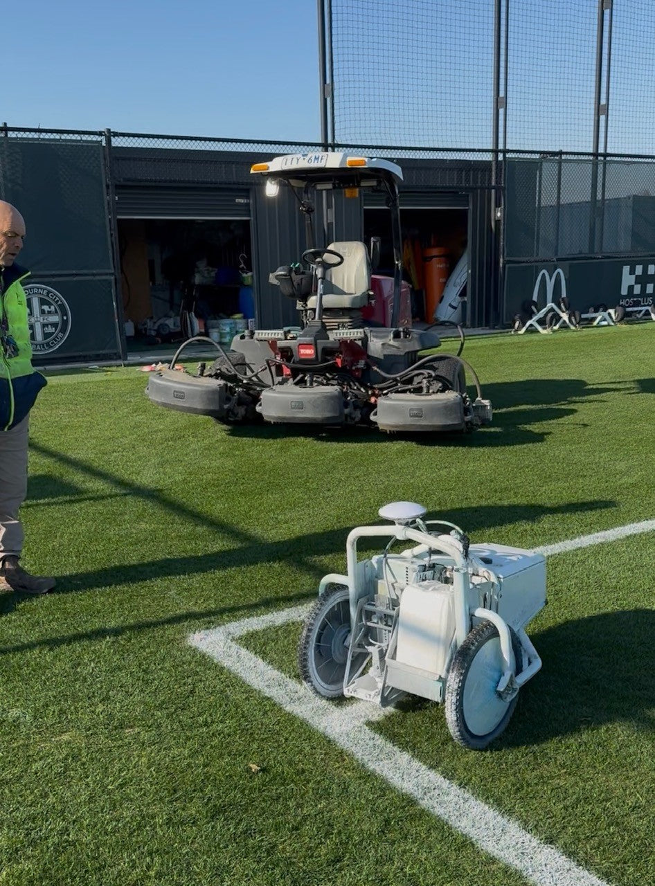 Robotic line marker beside freshly painted sports field lines, with a mower in the background
